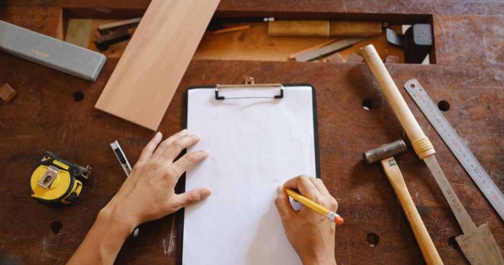 A hand writing a repair list on a clipboard beside a tape measure, hammer and hand tools on a workbench.
